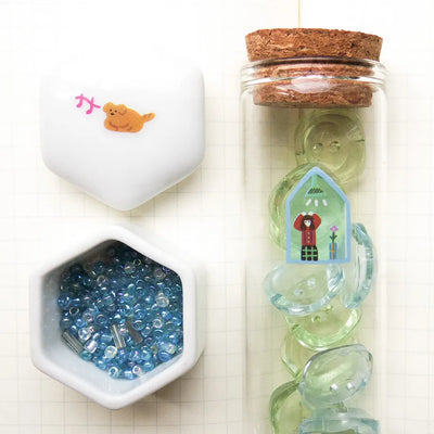 Decorative items including a small bottle with cork lid, a container with blue beads, and a hexagonal dish on a white background.