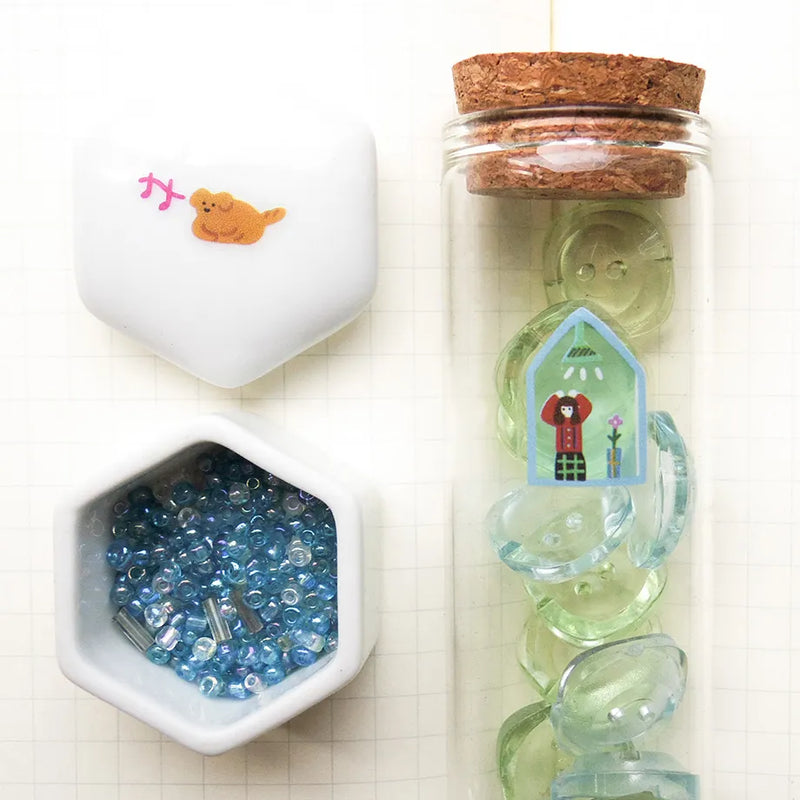 Decorative items including a small bottle with cork lid, a container with blue beads, and a hexagonal dish on a white background.