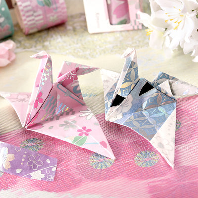 Decorative paper cranes on a pink fabric background with flowers and ribbons.