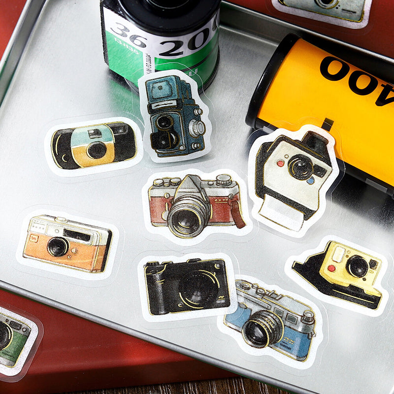 Collection of camera-shaped stickers on a metallic surface with film and camera equipment in the background.