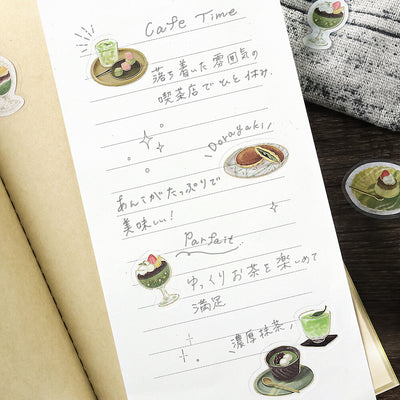 Sheet of paper with handwritten text and illustrations of tea and sweets on a wooden surface.