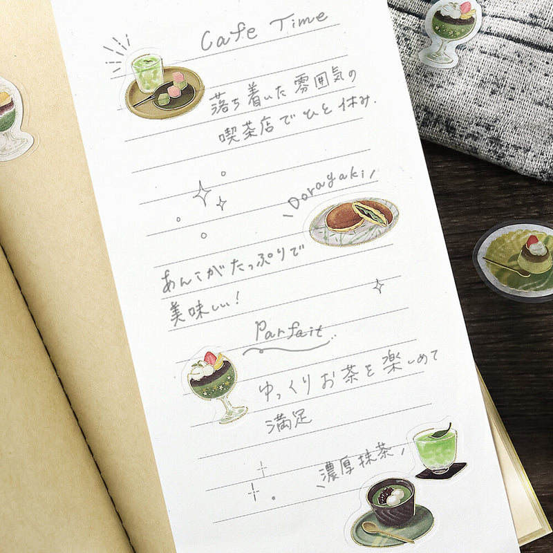 Sheet of paper with handwritten text and illustrations of tea and sweets on a wooden surface.
