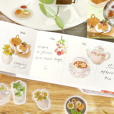 Weekly planner with illustrated stickers of tea, flowers, and food on a wooden surface.