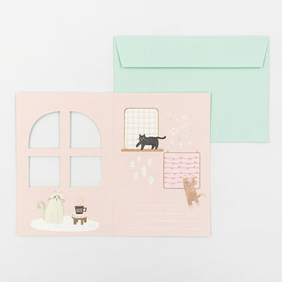 Illustrated scene with a cat, teapot, and window on a pink card with a green envelope.